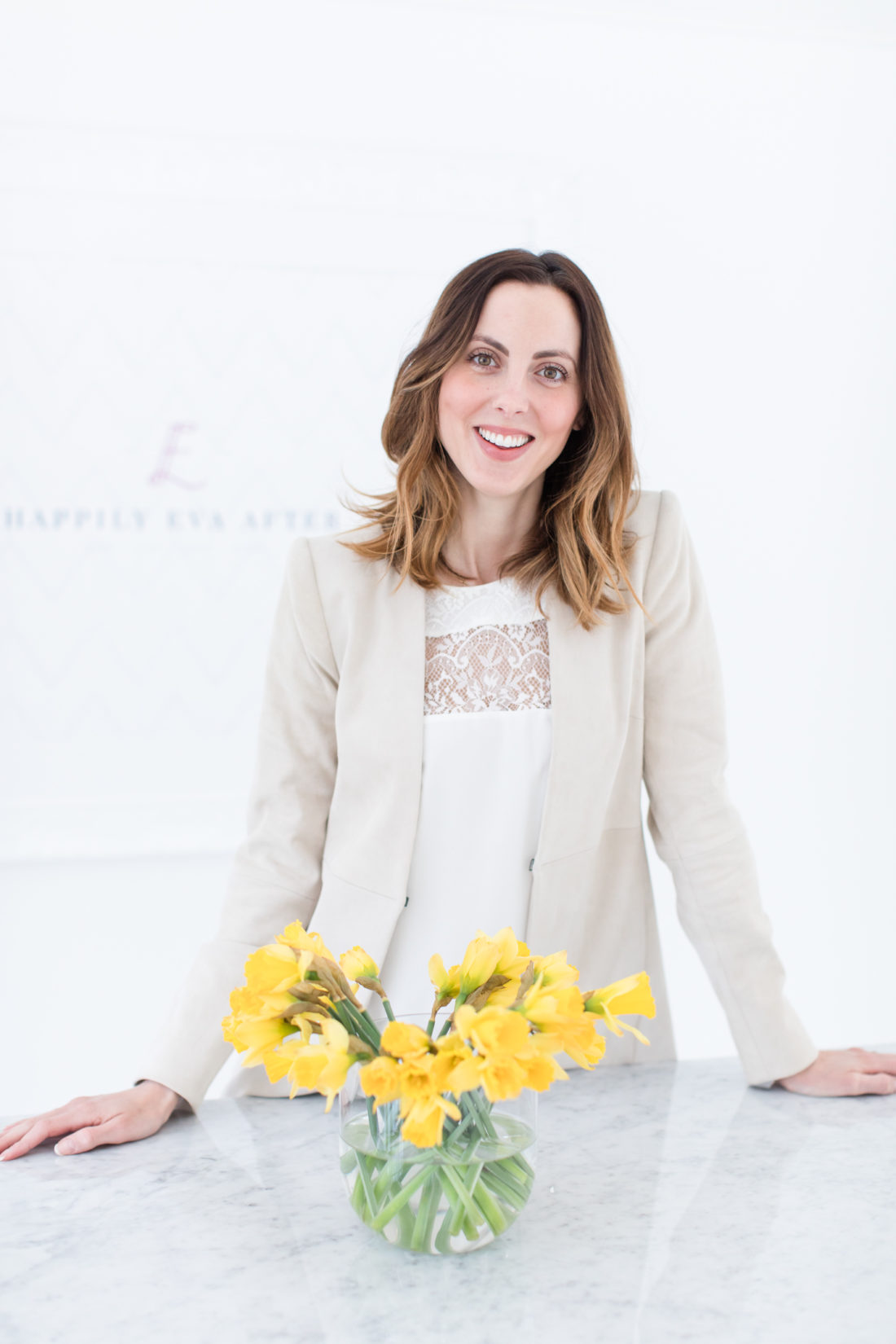Eva Amurri Martino wears a white camisole top and tan blazer, and stands in the happily eva after studio with a bowl of daffodils