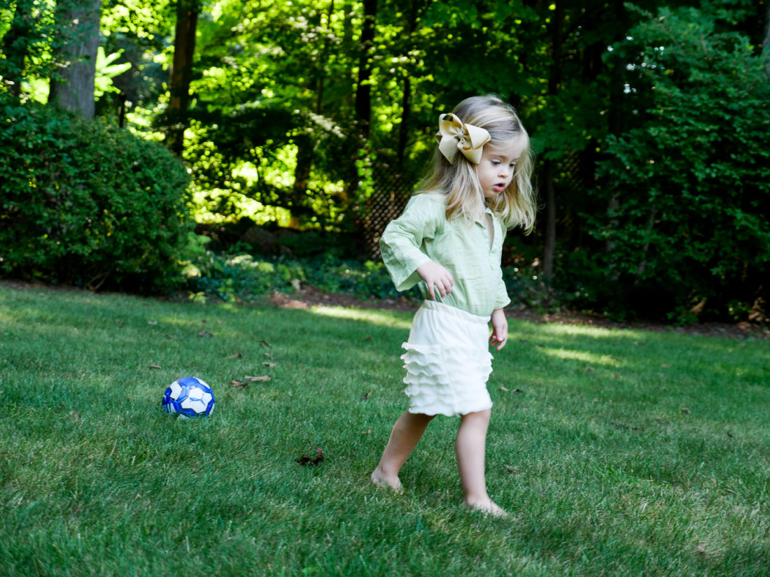 Marlowe Martino from the Happily Eva After Blog wearing a green tunic and ruffled white shorts and bow, walking on the grass at her home in Connecticut with a blue and white soccer ball