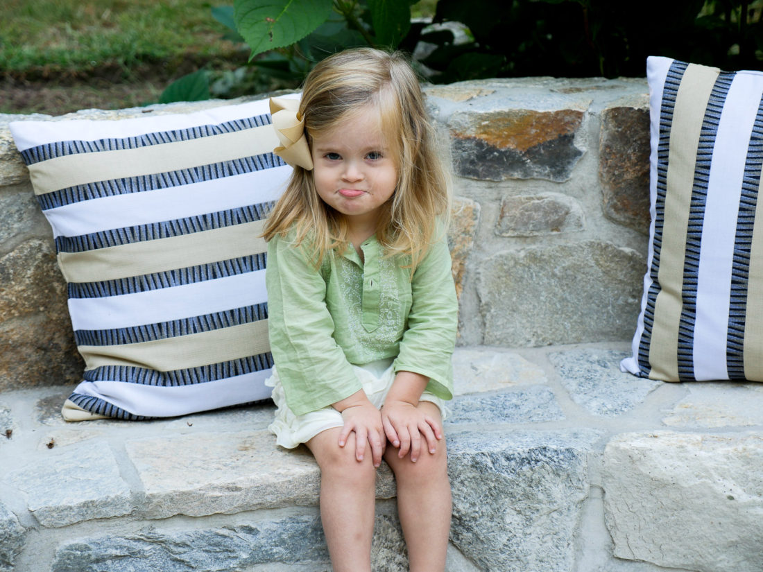 Marlowe Martino in a green tunic and white frilled shorts sitting outdoors on a stone bench with striped pillows