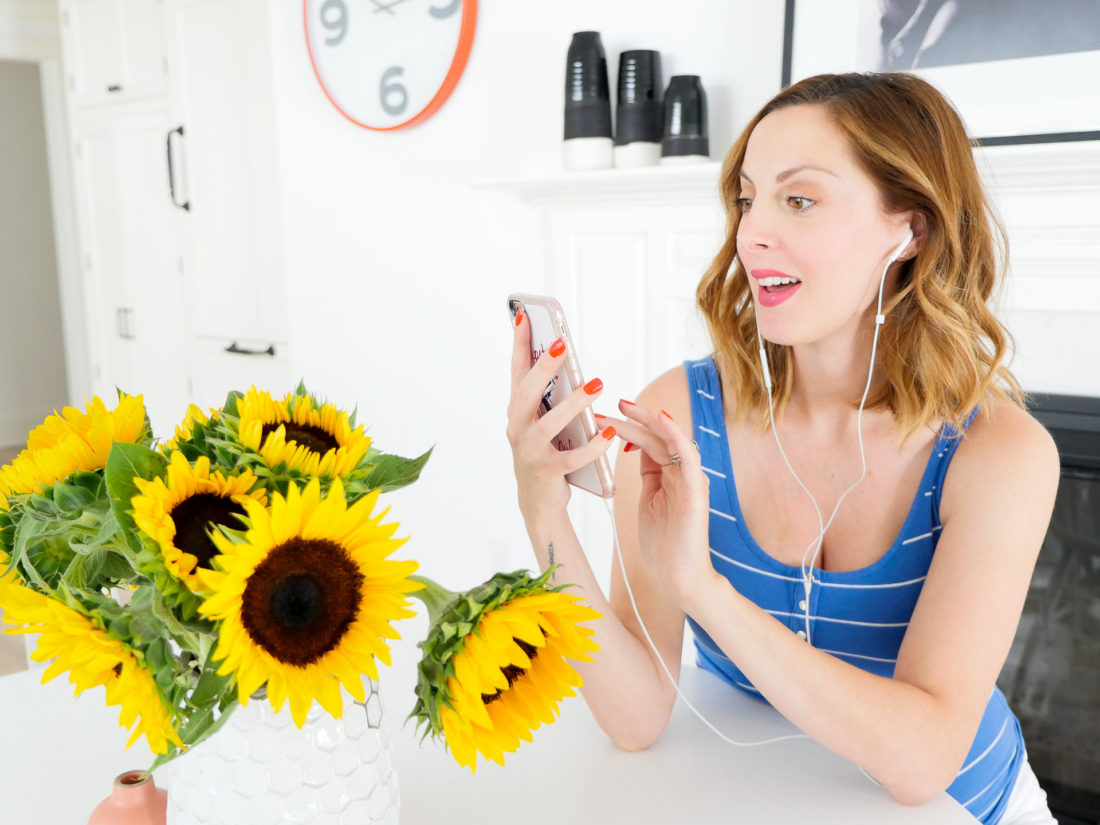 Eva Amurri Martino of blog Happily Eva After sitting at her kitchen table next to a vase of sunflowers, wearing a striped blue and white maternity tank and listening to an audiobook on Audible