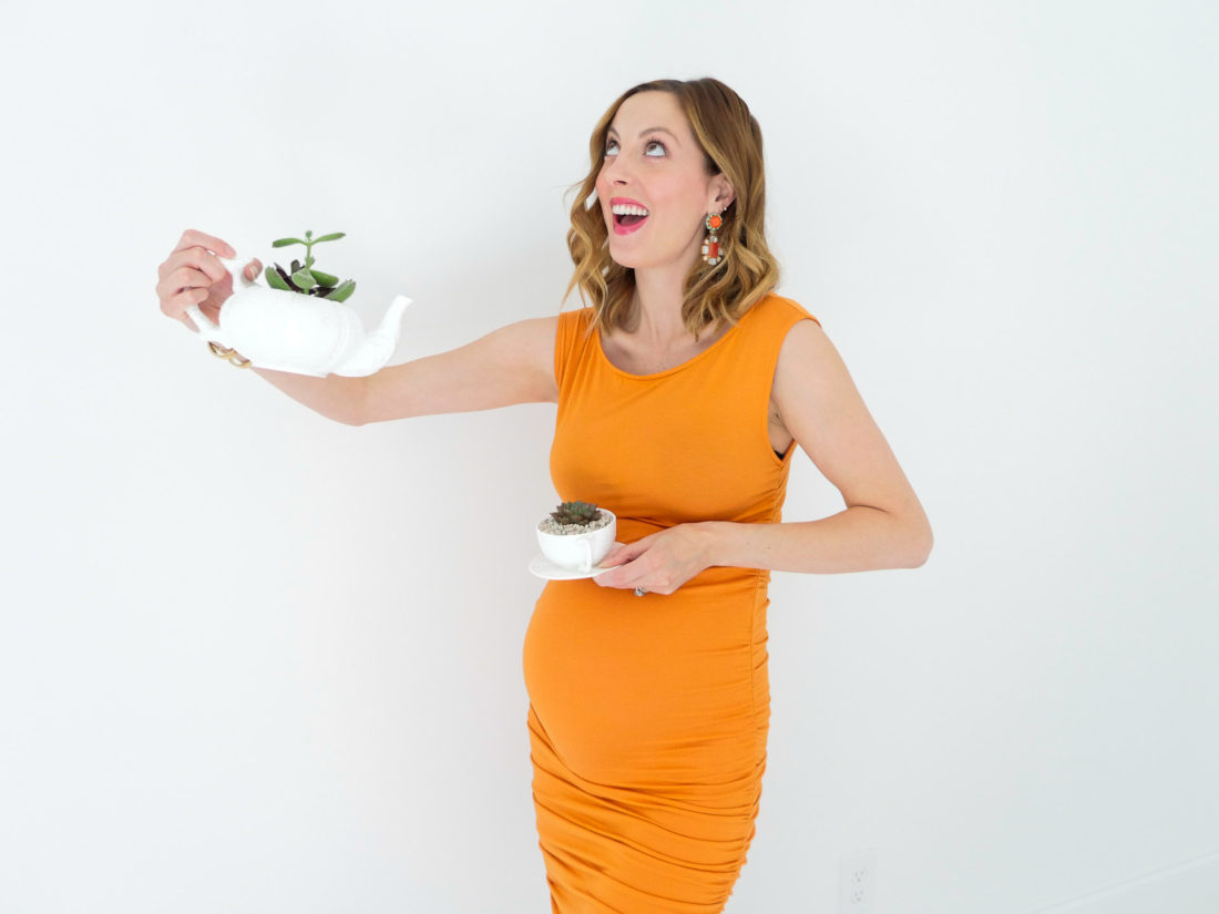 Eva Amurri Martino from the Happily Eva After blog displaying her DIY Teacup Planters with Mini Succulents and small pebbles, wearing a bodycon orange maternity dress, at thirty three weeks pregnant, colorful chandelier earrings, and gold bracelet watch