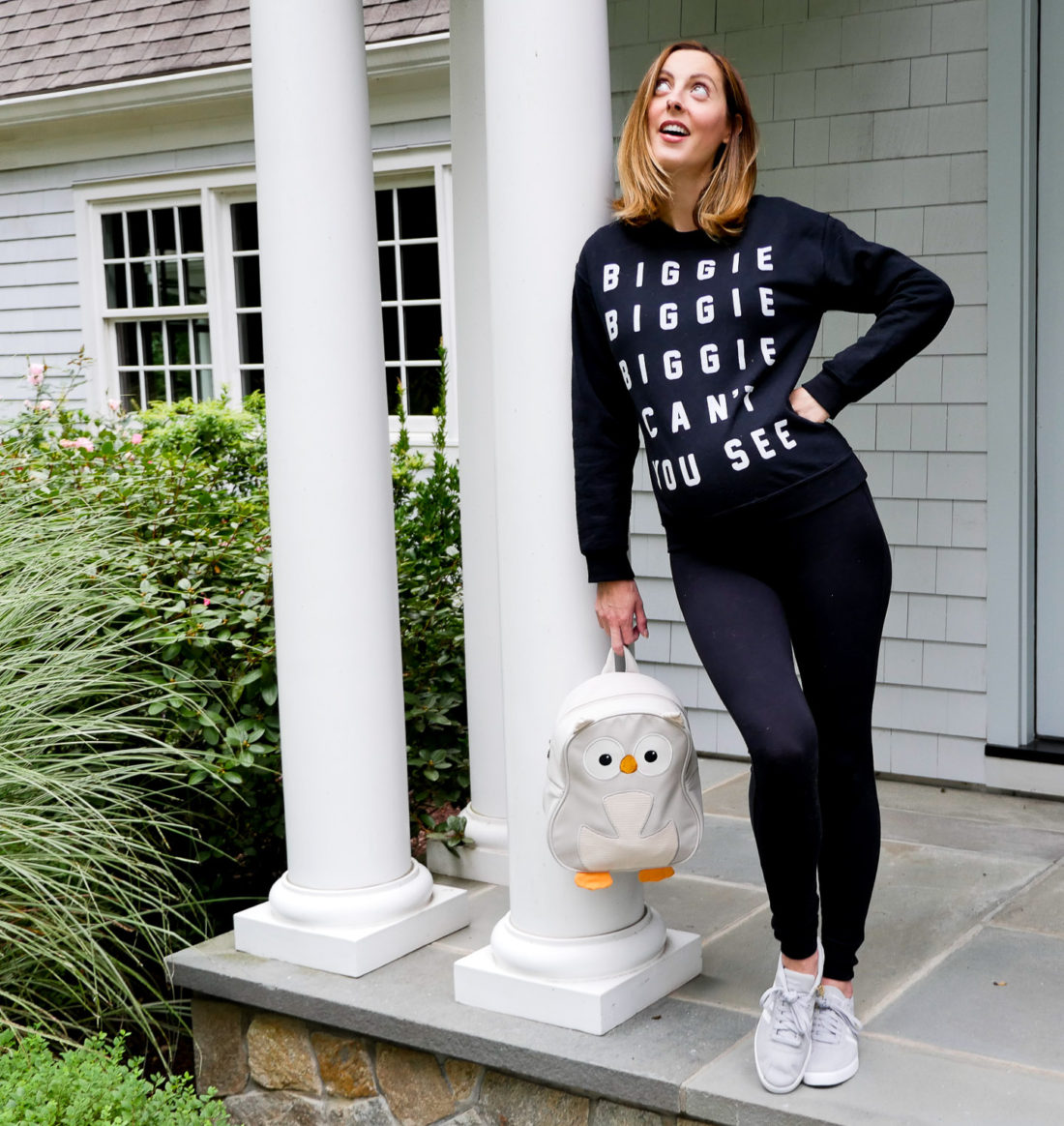 Eva Amurri Martino of Lifestyle blog Happily Eva After at 34 weeks pregnant wearing black maternity leggings and a "biggie biggie biggie can't you see" sweatshirt and addidas neo courtset sneakers holding her daughter Marlowe's light grey owl backpack