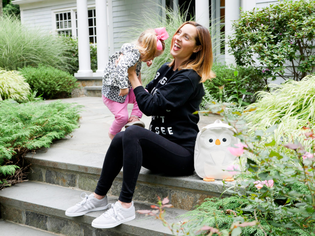 Eva Amurri Martino of lifestyle blog Happily Eva After sitting on the steps of her connecticut home wearing black maternity leggings, a blac sweatshirt, and light grey adidas sneakers, lifting her daughter Marlowe on to her lap