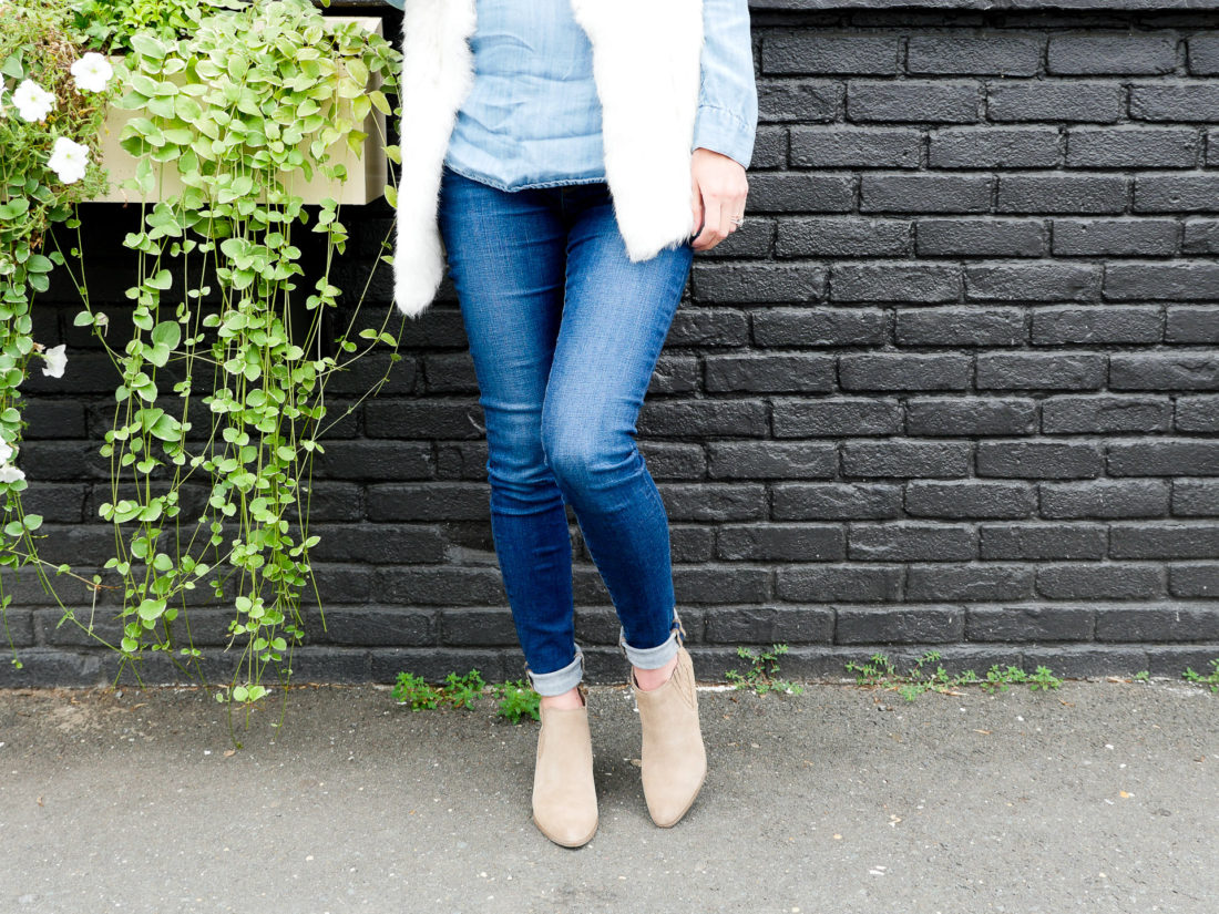 Eva Amurri Martino of Happily Eva After blog wearing maternity jeans, a chambray maternity top, a white fur vest, and tan colored fall boots standing against a dark grey wall