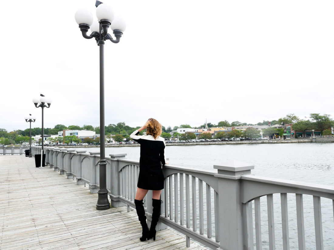 Eva Amurri Martino of lifestyle blog Happily Eva After wearing a mod black and cream shift dress and black over the knee boots facing out towards the water next to a lamp post