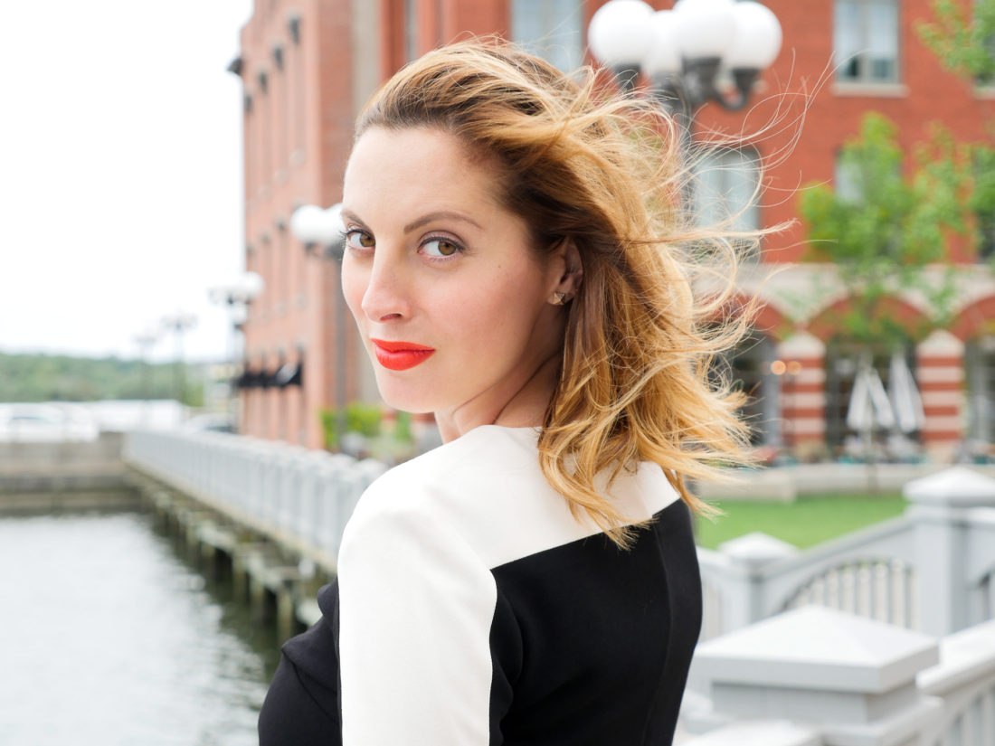 Eva Amurri Martino of lifestyle blog Happily Eva After wearing red lipstick and a cream and black dress standing at the waterfront in connecticut and looking back over her shoulder at the camera