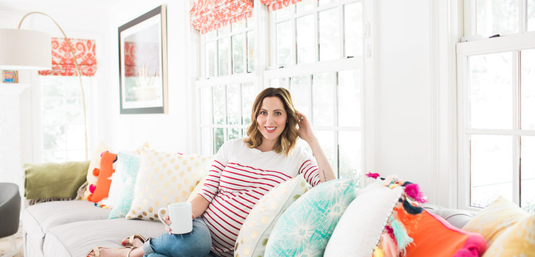 Eva Amurri Martin of lifestyle blog Happily Eva After relaxes at home in her family room on a grey couch with multicolored and multipatterned pillows surrounding her, holding a cup of coffee