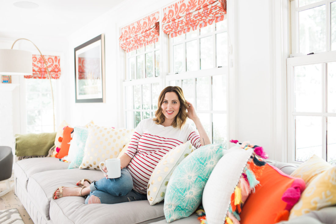 Eva Amurri Martin of lifestyle blog Happily Eva After relaxes at home in her family room on a grey couch with multicolored and multipatterned pillows surrounding her, holding a cup of coffee