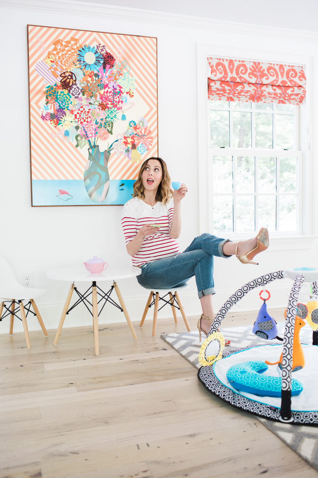 Eva Amurri Martin of lifestyle blog Happily Eva After drinking tea from her daughter's tea set and perching on a tiny white midcentury modern playroom furniture set with the jonathan adler crafter by fisher price collection sensory gym in the foreground