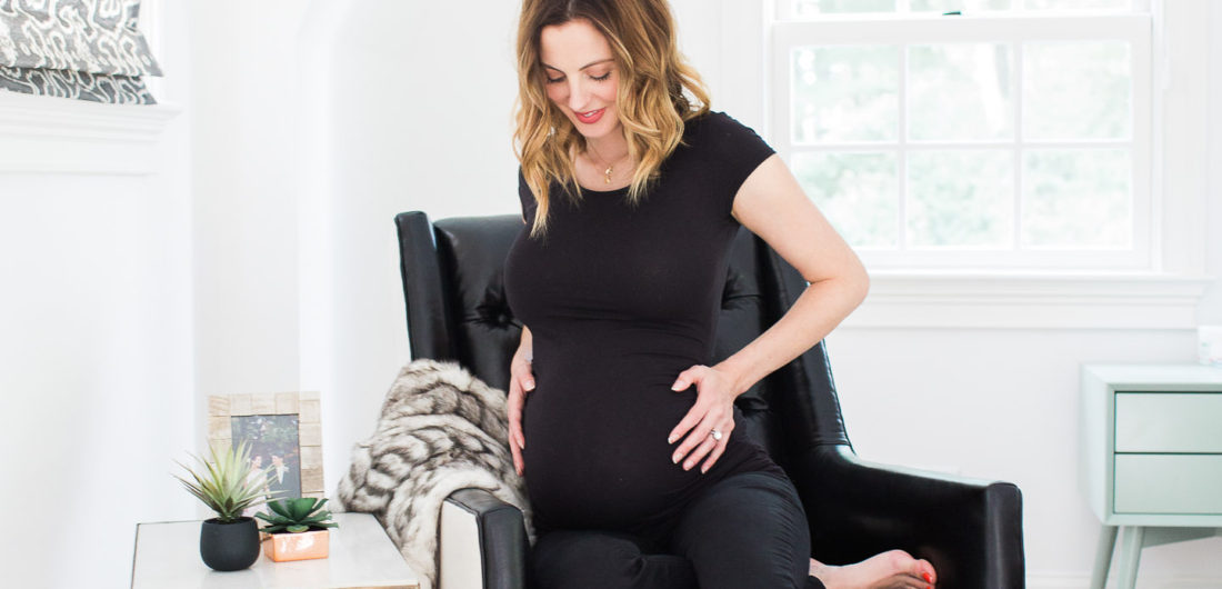 Eva Amurri Martino of lifestyle blog Happily Eva After cradling her 37 week bump while sitting in a black leather chair in her master bedroom next to a brass table and two succulents