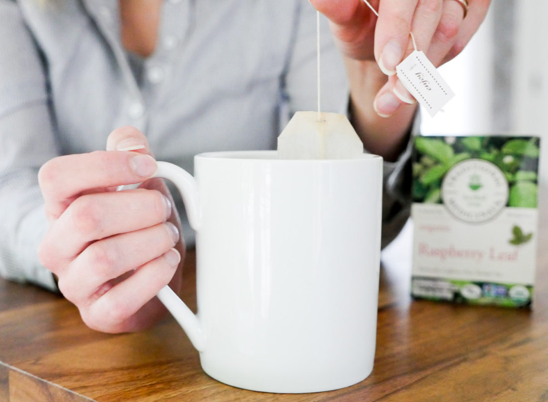 Eva Amurri Martino of lifestyle blog Happily Eva After sits at her dining room table in connecticut wearing a blue and white striped maternity top and dunking a tea bag in to a white mug