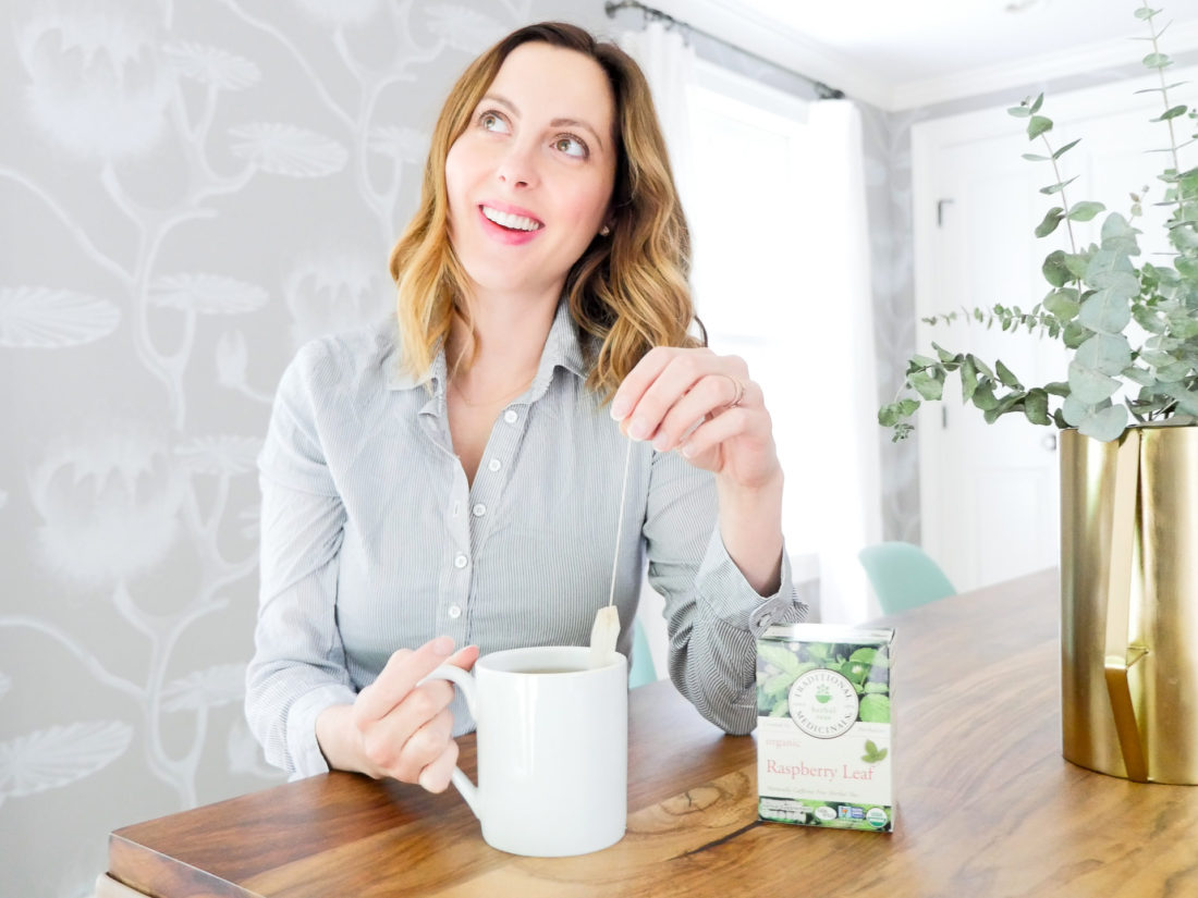 Eva Amurri Martino of lifestyle blog Happily Eva After sits at her dining room table in connecticut wearing a blue and white striped maternity top and dunking a tea bag in to a white mug