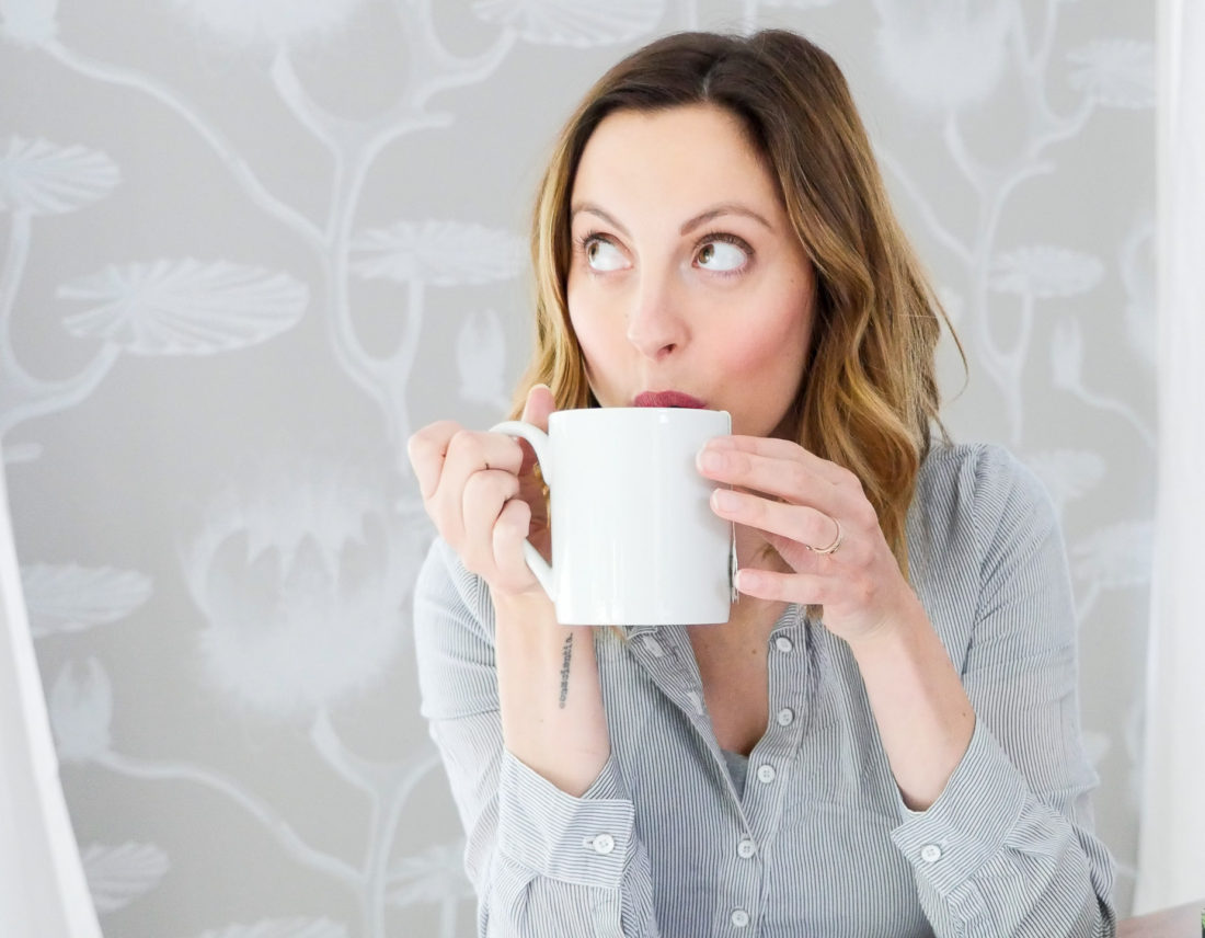 Eva Amurri Martino of lifestyle blog Happily Eva After sipping hot tea from a white mug while sitting in her dining room with a backdrop of grey and white patterned wallpaper