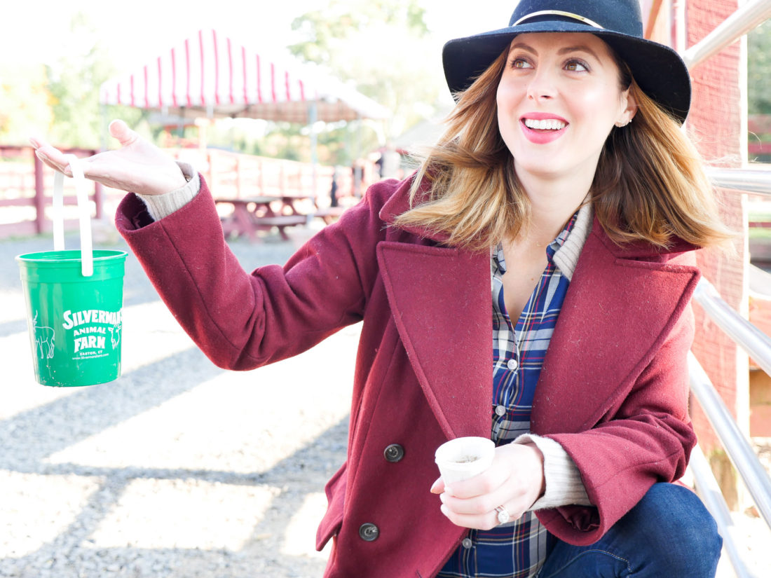 Eva Amurri Martino of lifestyle and motherhood blog Happily Eva After, wearing a plaid shirt and oxblood wool peacoat, holding a feed bucket at Silverman's farm in connecticut