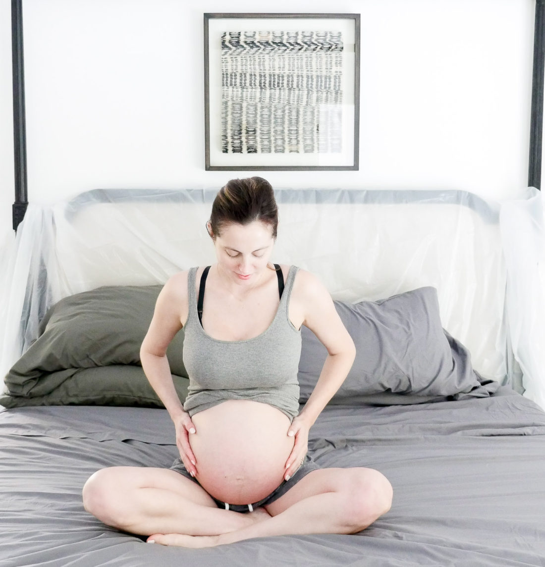 Eva Amurri Martino of lifestyle and motherhood blog Happily Eva After in the early stages of labor at her home in connecticut, sitting on her bed in a grey tank top and grey shorts cradling her baby bump