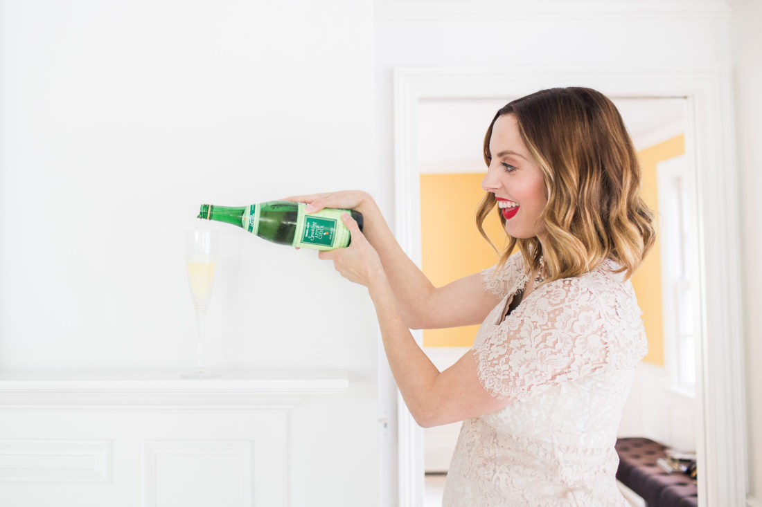 Eva Amurri Martino of lifestyle blog Happily Eva After wearing a champagne colored lace maternity dress and pouring a champagne glass full of sparkling cider