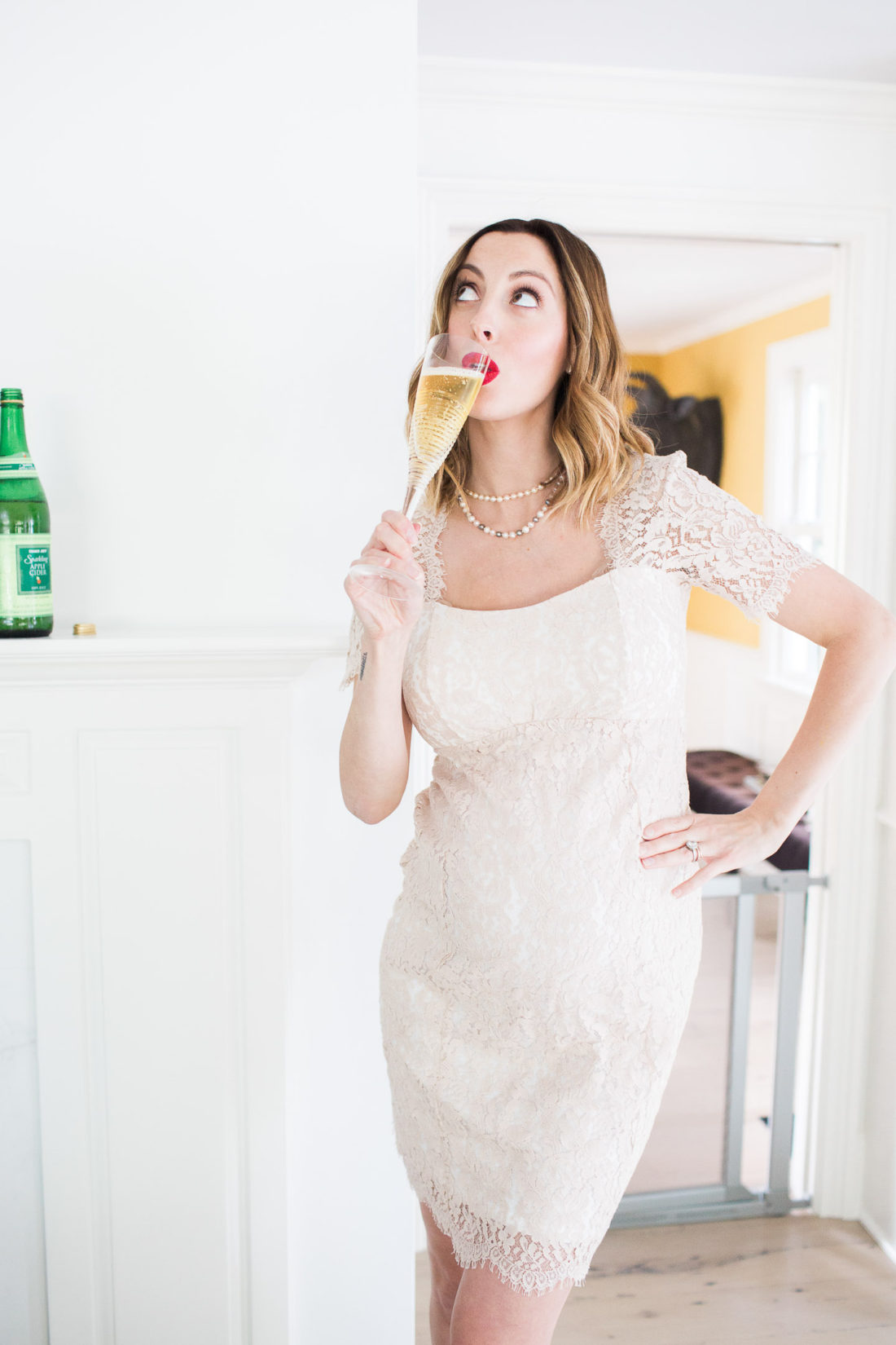 Eva Amurri Martino of lifestyle and motherhood blog Happily Eva After, standing in a champagne colored lace dress and red louboutin platform pumps, holding a glass of sparkling cider in a celebratory toast