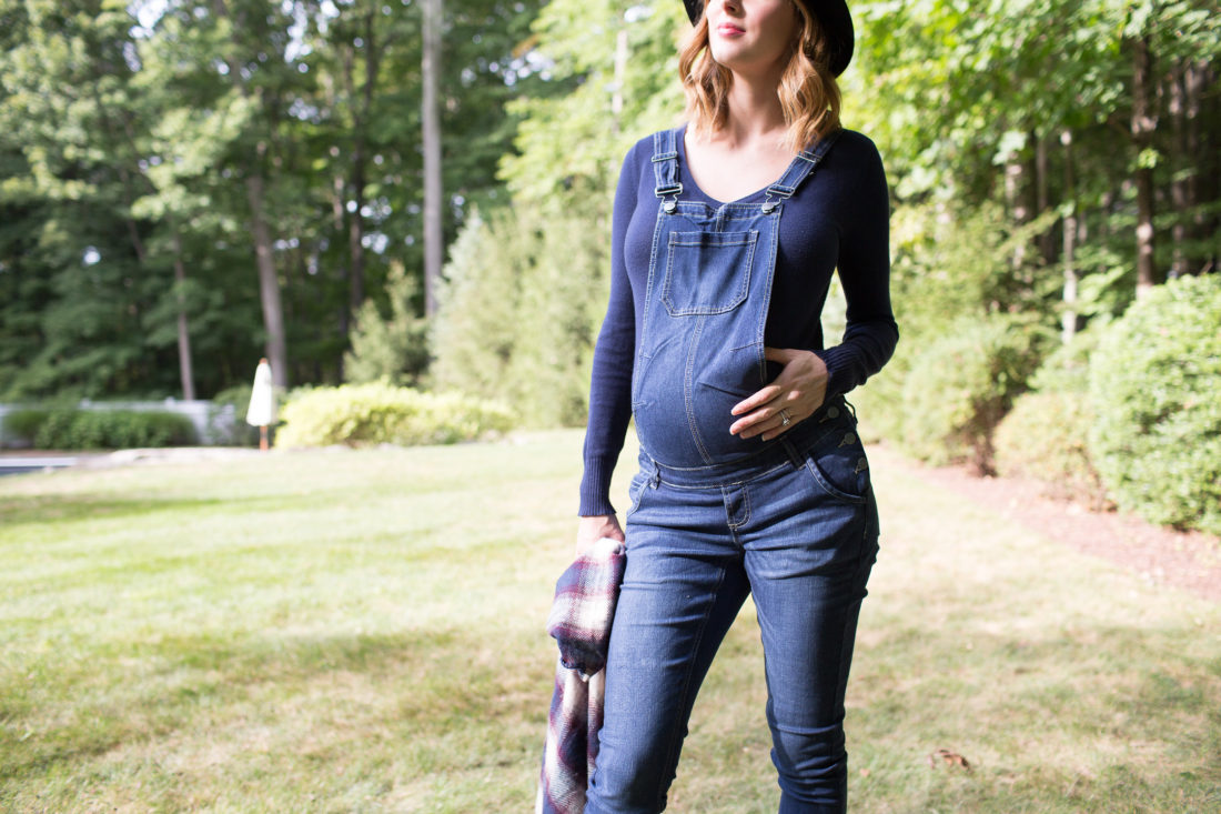 Eva Amurri Martino of lifestyle blog Happily Eva After standing in her yard in maternity overalls, a navy felt hat, and a plaid poncho at her home in connecticut