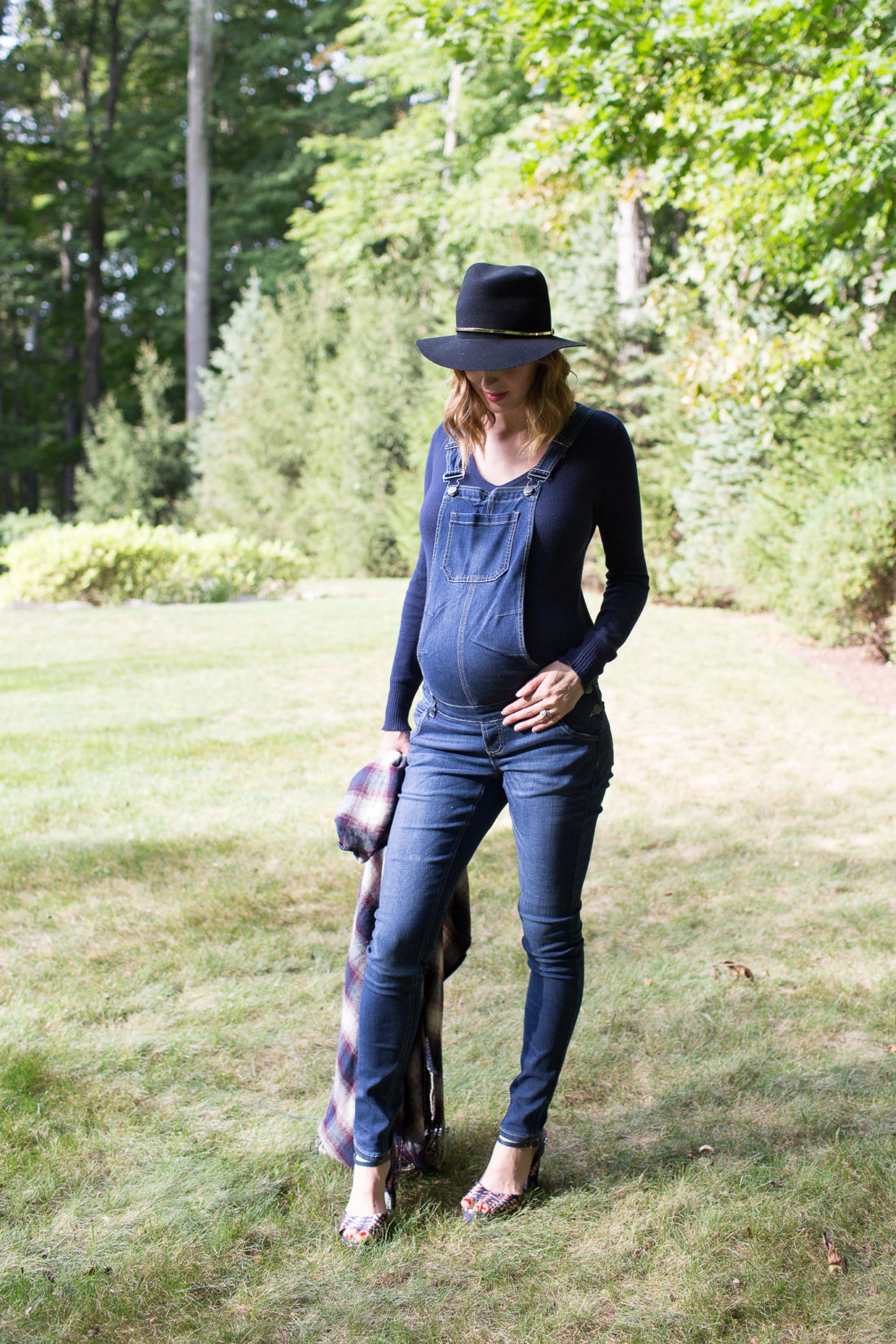 Eva Amurri Martino of lifestyle blog Happily Eva After standing in her yard in maternity overalls, a navy felt hat, and a plaid poncho at her home in connecticut