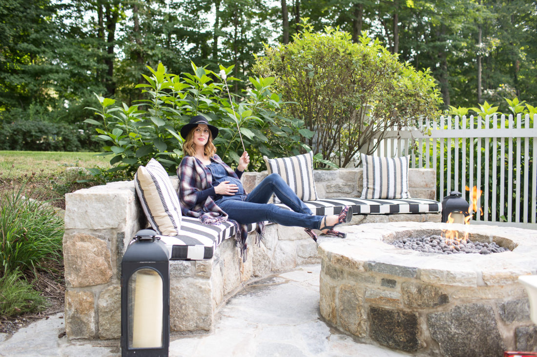 Eva Amurri Martino of lifestye and motherhood blog Happily Eva After at her firepit at her connecticut home, wearing maternity overalls, a navy felt hat, and a plaid poncho roasting marshmallows over the fire