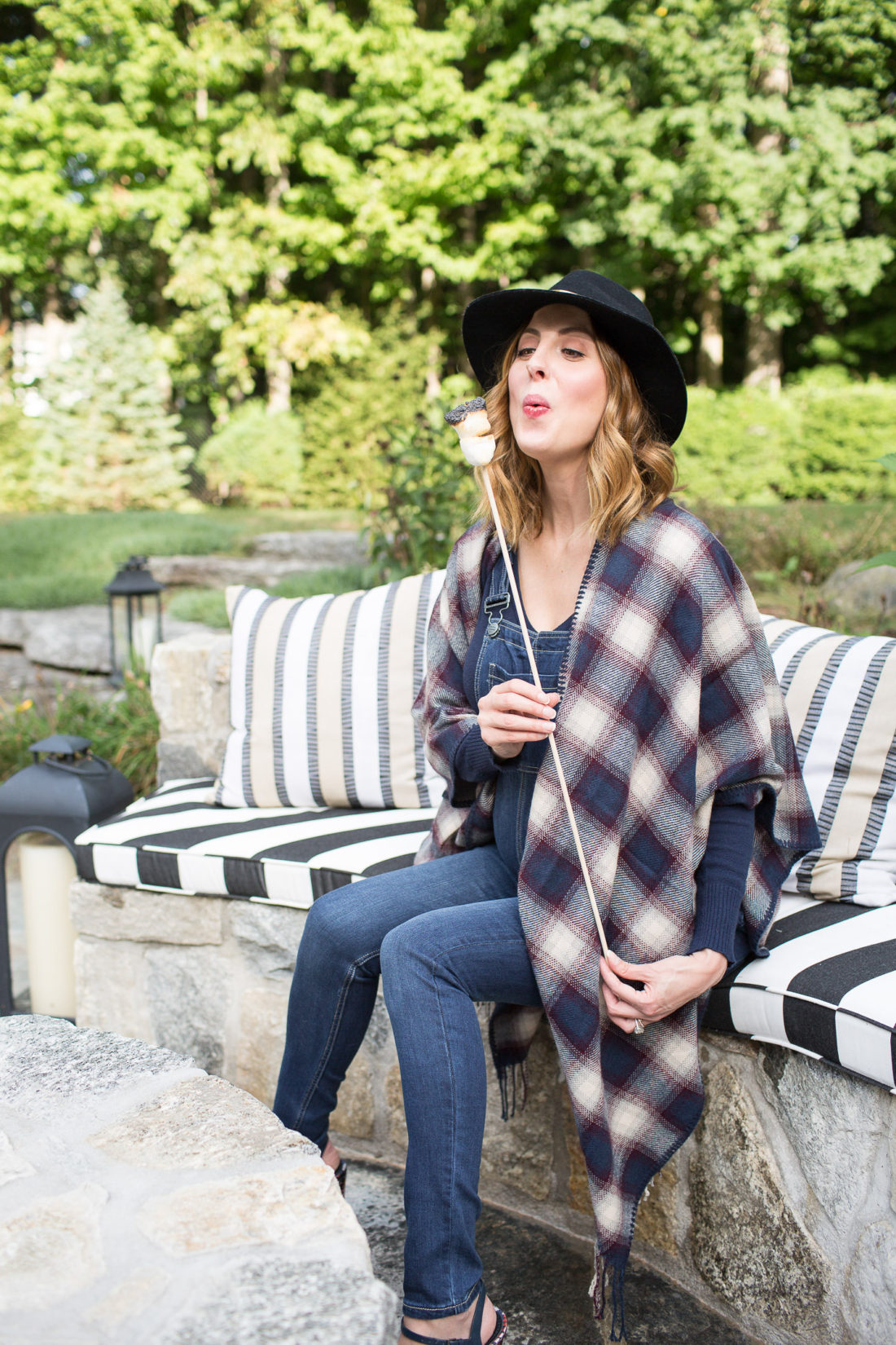 Eva Amurri Martino of lifestye and motherhood blog Happily Eva After at her firepit at her connecticut home, wearing maternity overalls, a navy felt hat, and a plaid poncho roasting marshmallows over the fire