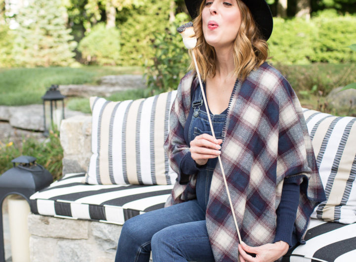 Eva Amurri Martino of lifestye and motherhood blog Happily Eva After at her firepit at her connecticut home, wearing maternity overalls, a navy felt hat, and a plaid poncho roasting marshmallows over the fire