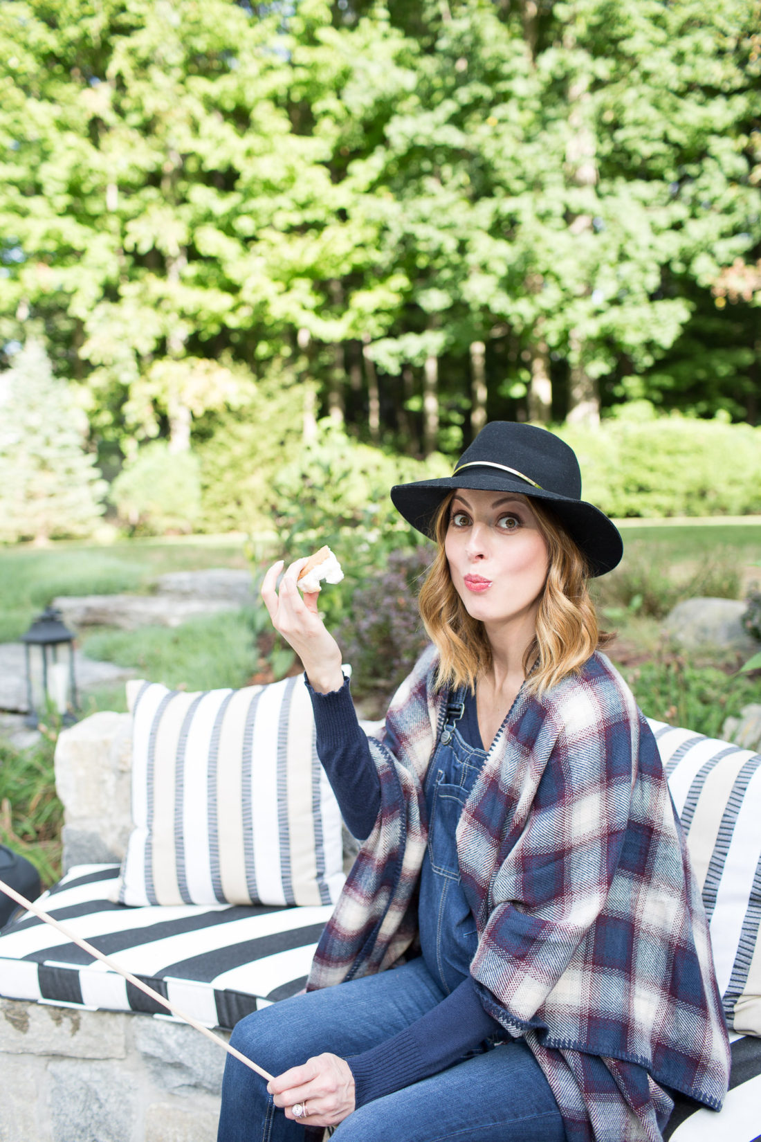 Eva Amurri Martino of lifestyle and motherhood blog Happily Eva After biting in to a S'more at her fire pit while wearing a plaid poncho and navy blue felt hat at 38 weeks pregnant