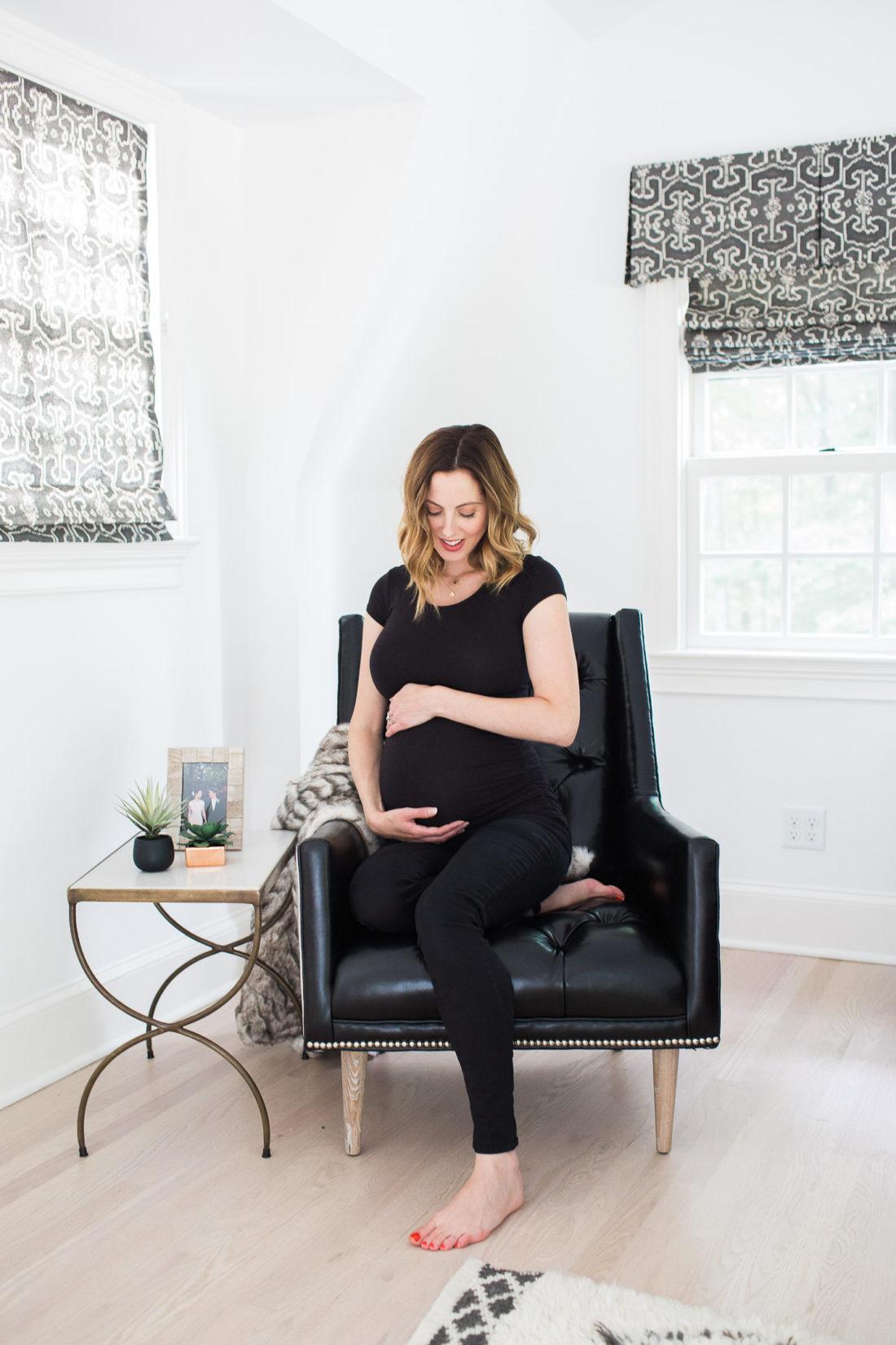 Eva Amurri Martino of lifestyle and motherhood blog Happily Eva After cradling her baby bump in a black maternity top and jeans while sitting on a leather armchair in her master bedroom