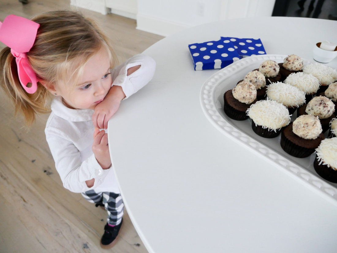 Marlowe Martino wearing a white top and pink bow, eyeing a tray of cupcakes