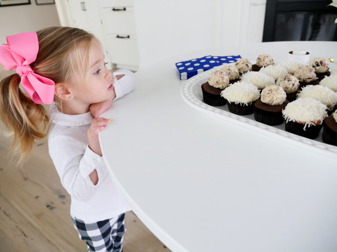 Marlowe Martino wearing a white shirt and big pink bow, eyeing a tray of cupcakes