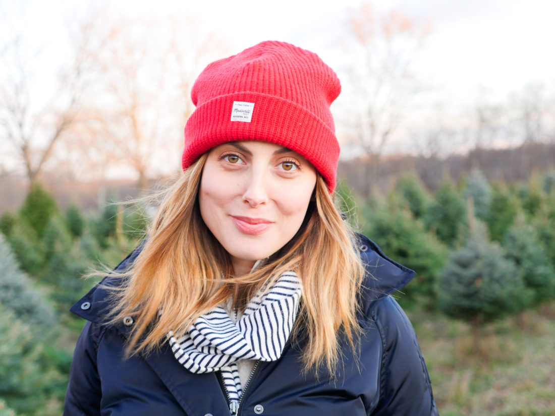 Eva Amurri Martino of lifestyle and motherhood blog Happily Eva After wears a navy puffer jacket, blue and white striped scarf and red wool hat at the Christmas tree farm in Connecticut