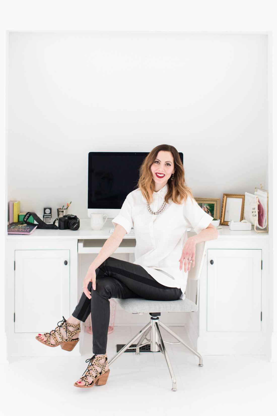 Eva Amurri Martino sits at her desk for blog Happily Eva After, wearing a white button down shirt, leather leggings, leopard print sandals, and red lipstick