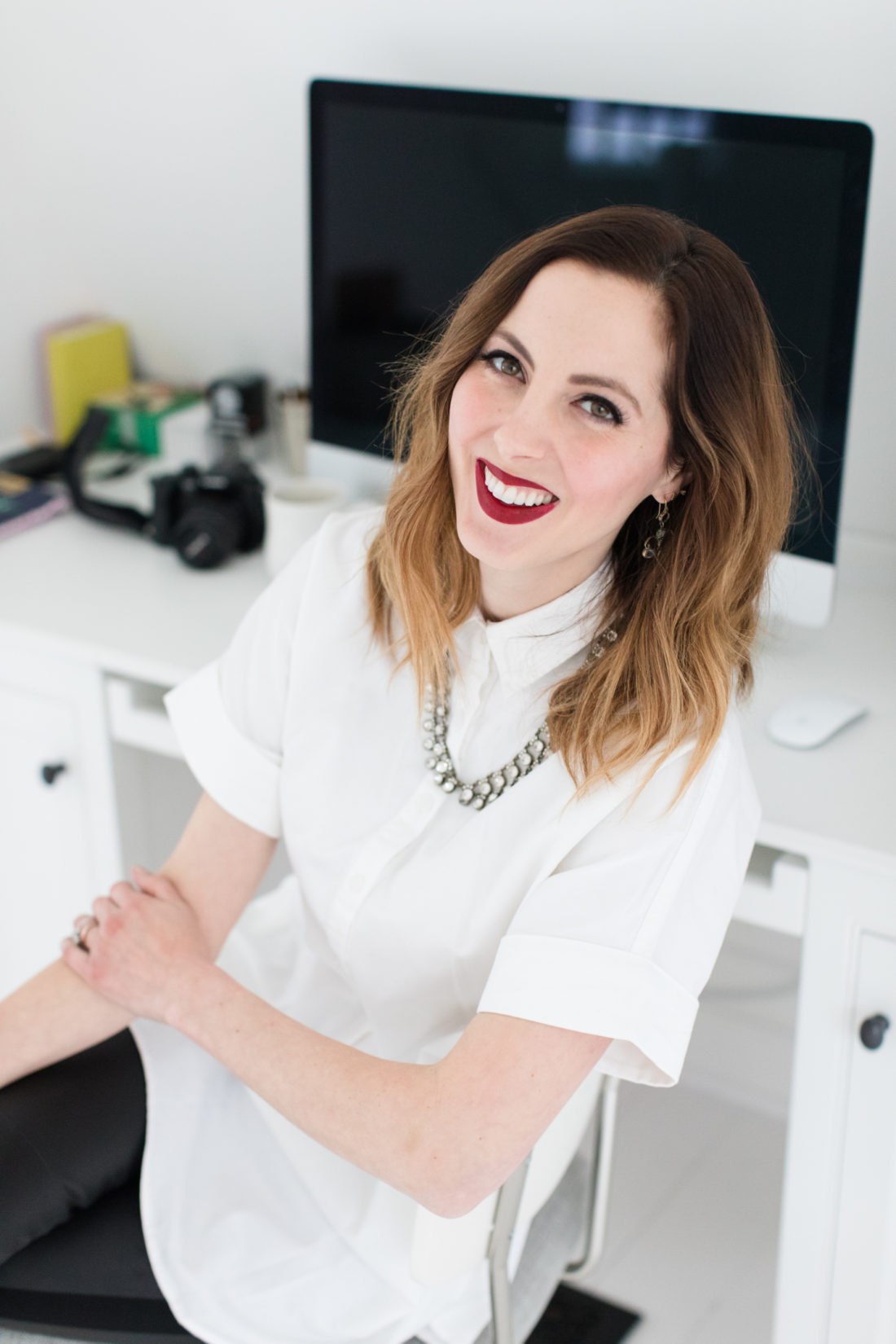 Eva Amurri Martino of lifestlye and motherhood blog Happily Eva After wears a crisp white button down shirt, a statement necklace, and red lipstick at her desk in Connecticut