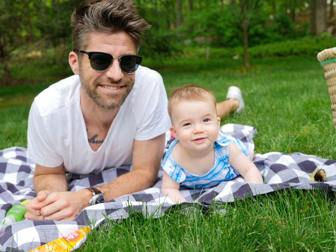 Kyle Martino relaxes on a picnic basket wearing sunglasses and a white teeshirt with seven month old son, Major