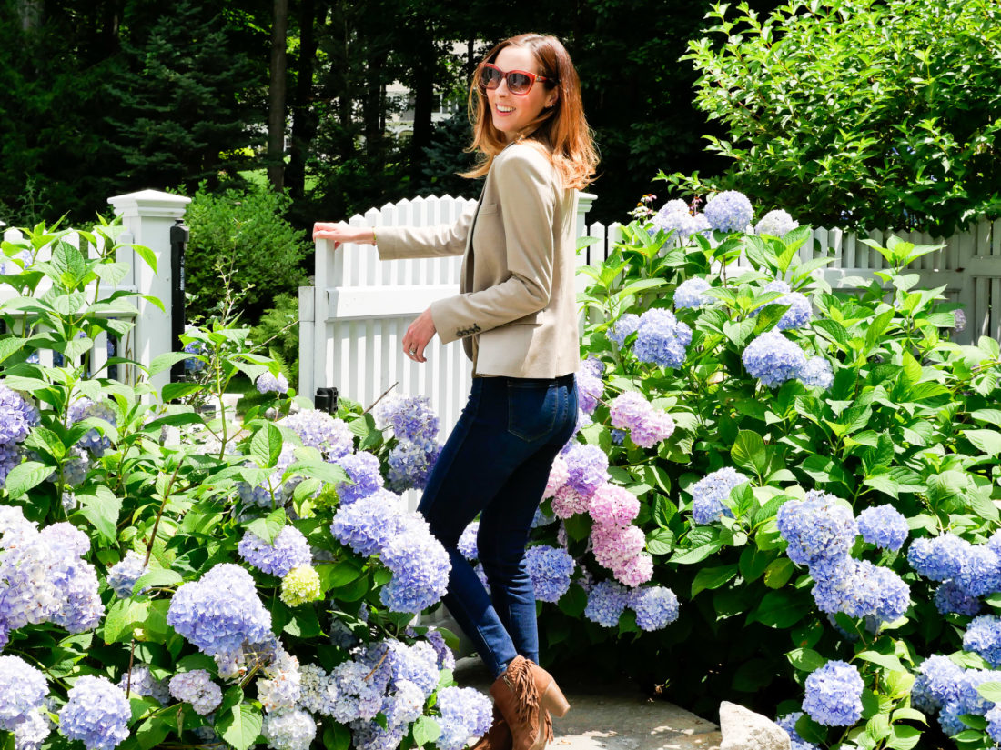 Eva Amurri Martino wears dark denim, brown fringe booties, an olive green sweater and blazer in the garden of her Connecticut home