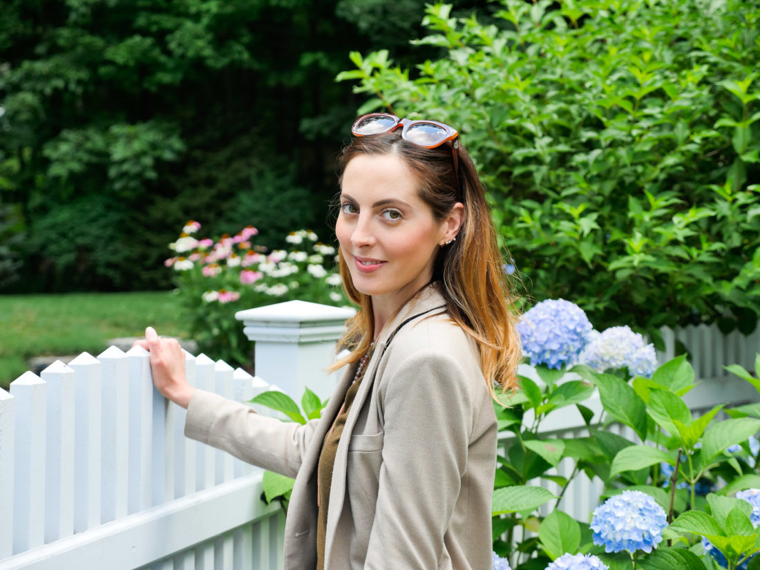 Eva Amurri Martino wears dark denim, brown fringe booties, an olive green sweater and blazer in the garden of her Connecticut home
