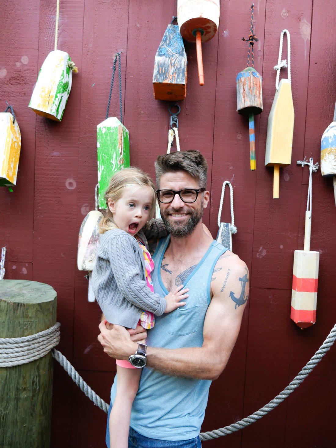 Kyle Martino and daughter Marlowe stand in front of a wall of multicolored buoys in downtown Bar Harbor, ME