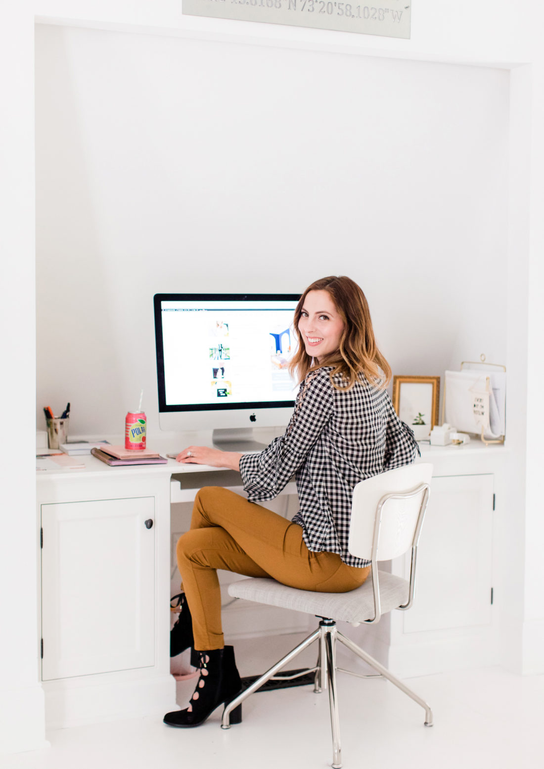 Eva Amurri Martino wears tan jeans and a black and white checked top, and sits at her computer in the Happily Eva After studio