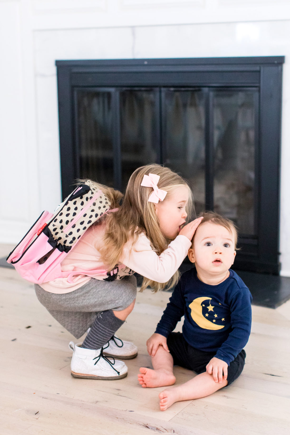 Marlowe Martino wears a pink sweatshirt, grey skirt, and kitty cat backpack as she leans down to pat little brother Major on the head before she heads off to school
