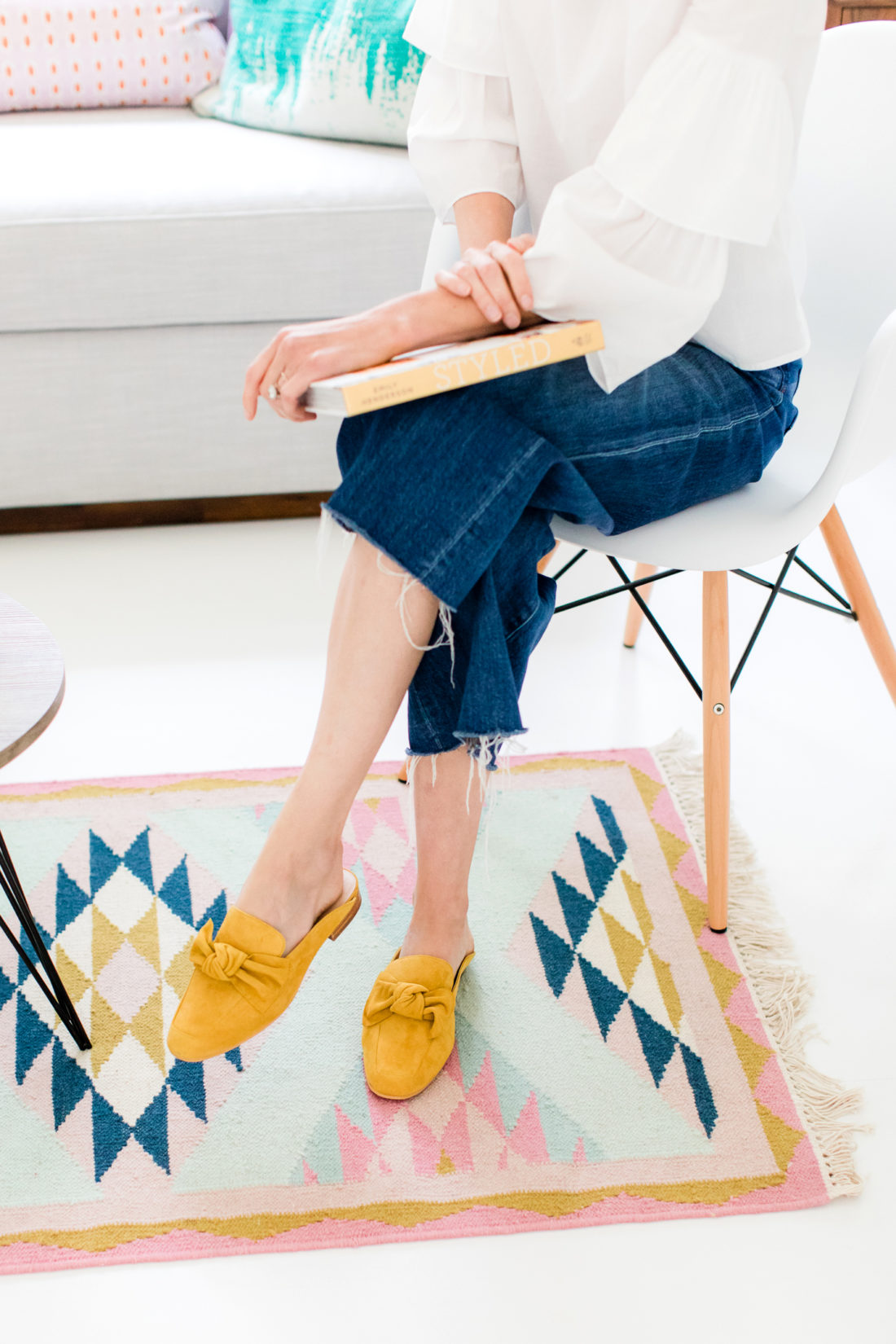 Eva Amurri Martino relaxes in her home office reading a book, wearing a white tiered top, frayed cropped denim, and mustard mules