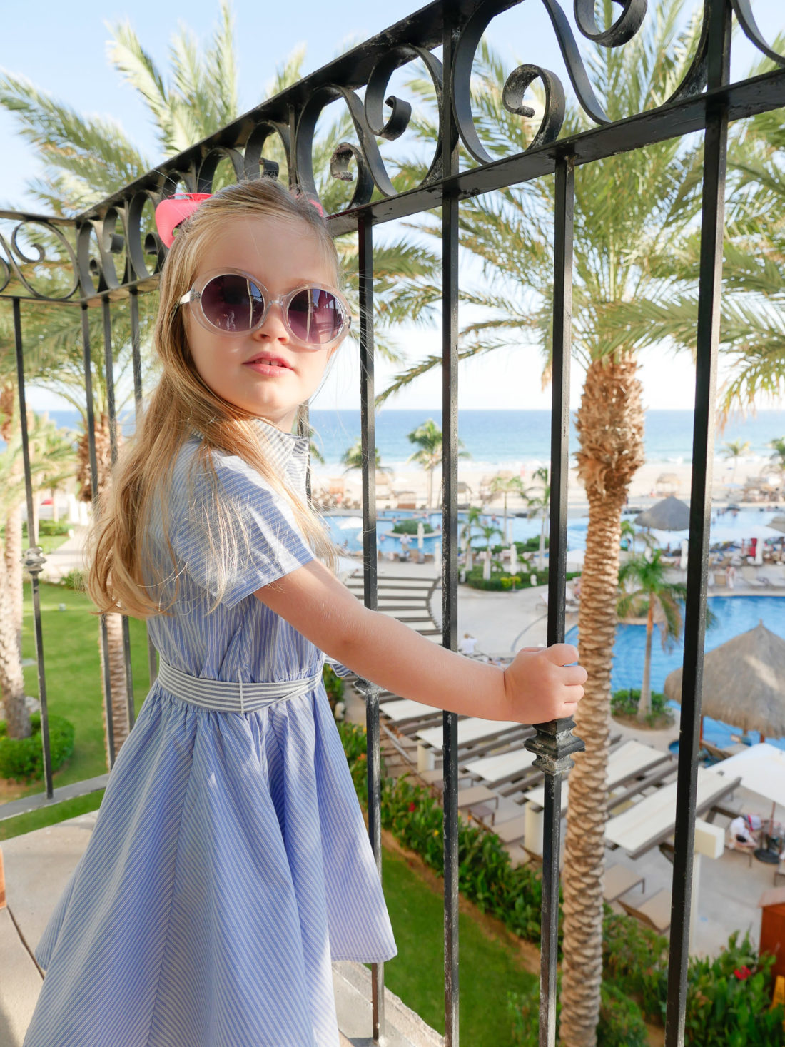 Marlowe Martino stands on the balcony wearing a blue and white dress and sunglasses at the Hyatt Ziva resort in Los Cabos Mexico
