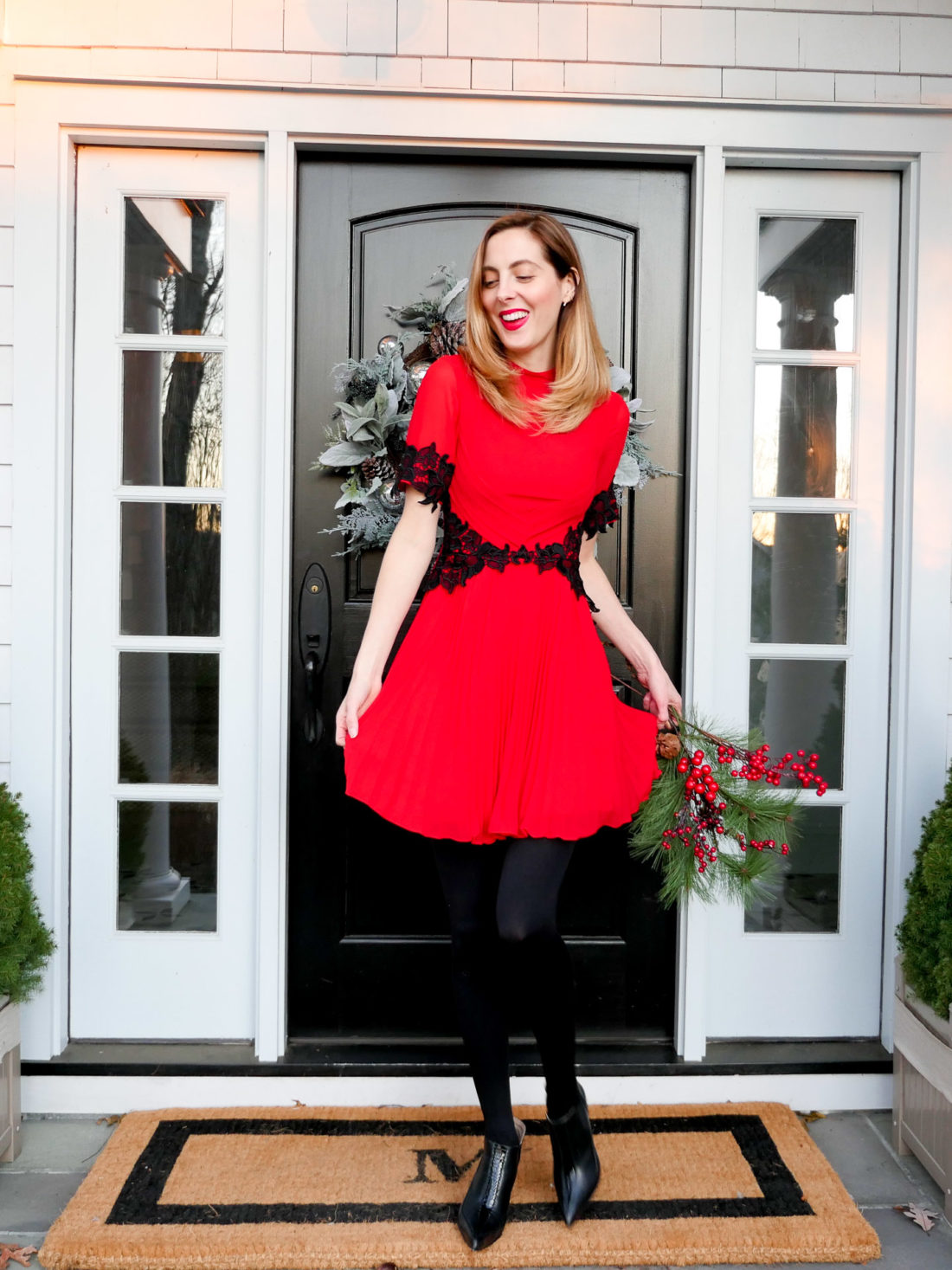Eva Amurri Martino wears a red holiday party dress with black lace detailing, and stands in front of her Connecticut home