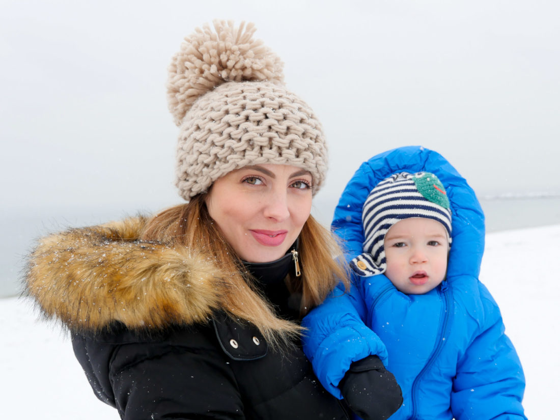 Eva Amurri Martino wears a tan knit beanie and faux fur lined parka, and holds one year old son Major on the snowy beach in Westport, CT