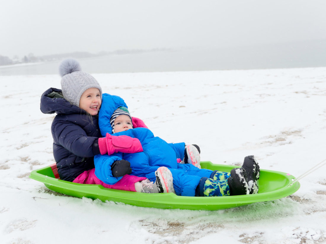 Marlowe Martino holds little brother Major in snowsuits on a green sled on the snowy beach in Westport, CT