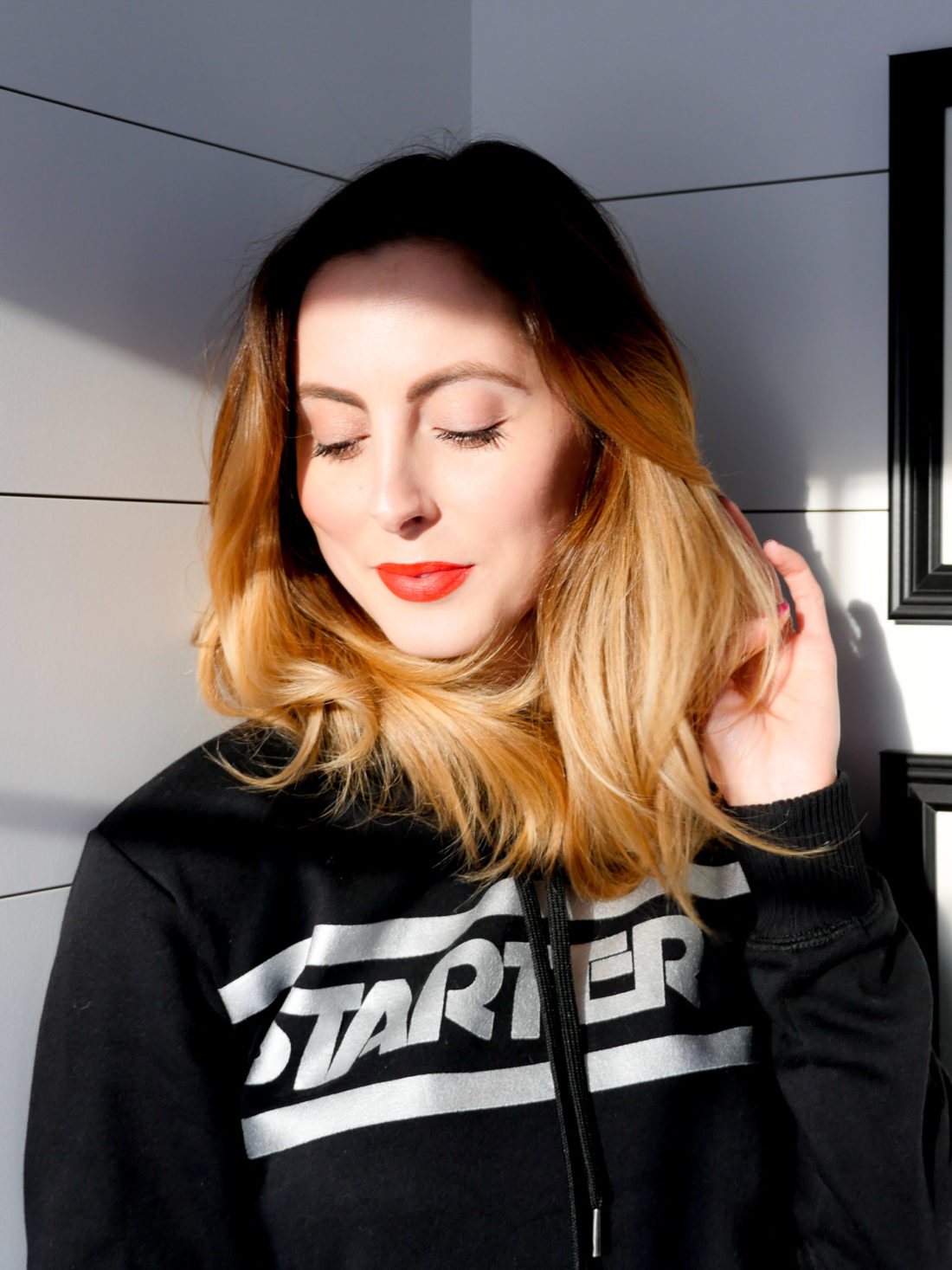 Eva Amurri Martino wears a Starter hoodie with red lipstick and stand in a beam of sunlight