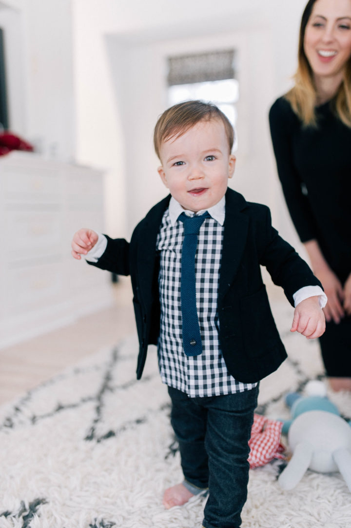 Major James Martino walking on the rug in his parents bedroom wearing a navy blue blazer and gingham shirt with a tie and jeans while his mother, Eva Amurri Martino, watches on from behind. 