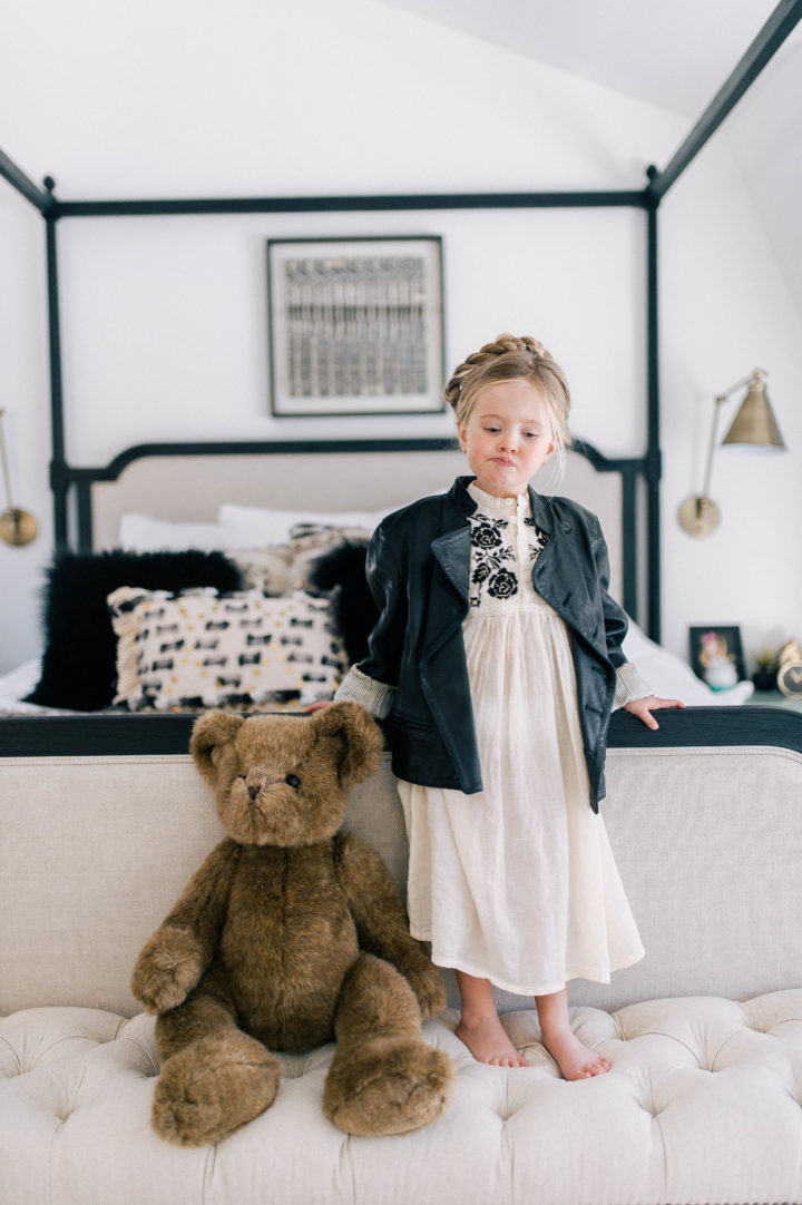 Marlowe Mae Martino pouting with a teddy bear, wearing an ivory tunic dress, leather jacket, and a halo braid in her parents bedroom in their Connecticut home.