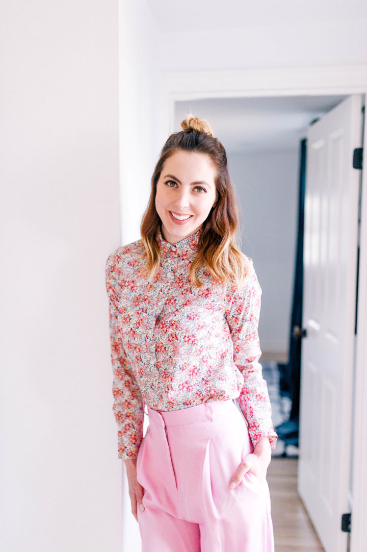 Eva Amurri Martino wears wide pink trousers, a pink floral button up shirt, and her hair in a half up half down topknot in her Connecticut home.