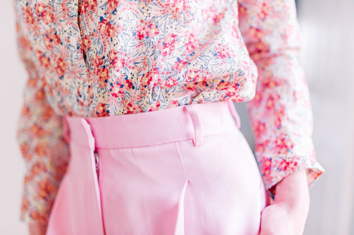 A closeup of Eva Amurri Martino wears wide pink trousers, a pink floral button up shirt, and her hair in a half up half down topknot in her Connecticut home.