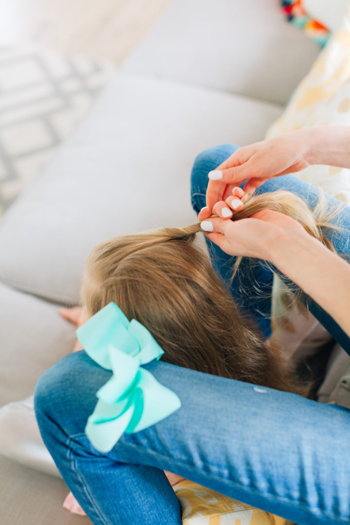 Eva Amurri Martino styles her daughter Marlowe's hair while she sits on the couch with a blue bow hairtie on her knee.